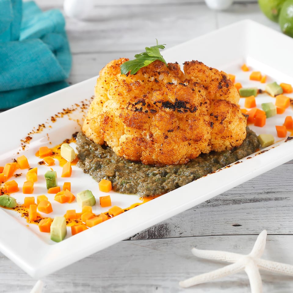 Roasted cauliflower steak on a bed of green sauce, garnished with diced carrots and avocado.