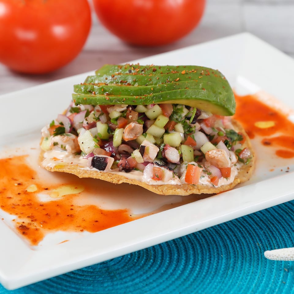 A tostada topped with ceviche, sliced avocado, and chili flakes on a white plate.