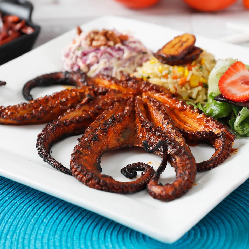 Grilled octopus with rice, coleslaw, salad with strawberries, and plantains on a white plate.