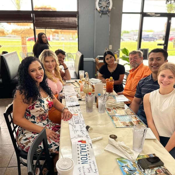 A diverse group of people smiling and sitting around a table with drinks and a "Greater Dalton Chamber" banner.