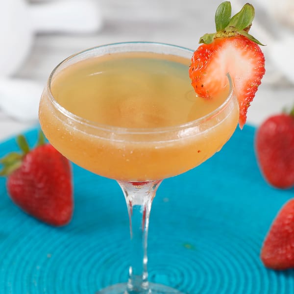 A coupe glass with a strawberry-garnished cocktail on a blue placemat with strawberries and starfish.