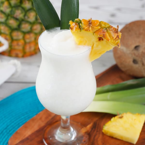 A frozen pina colada garnished with pineapple and leaves, next to a whole pineapple and coconut.