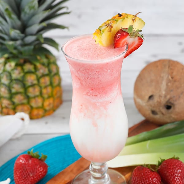 Pink and white layered tropical drink garnished with pineapple and strawberry, surrounded by fruit.